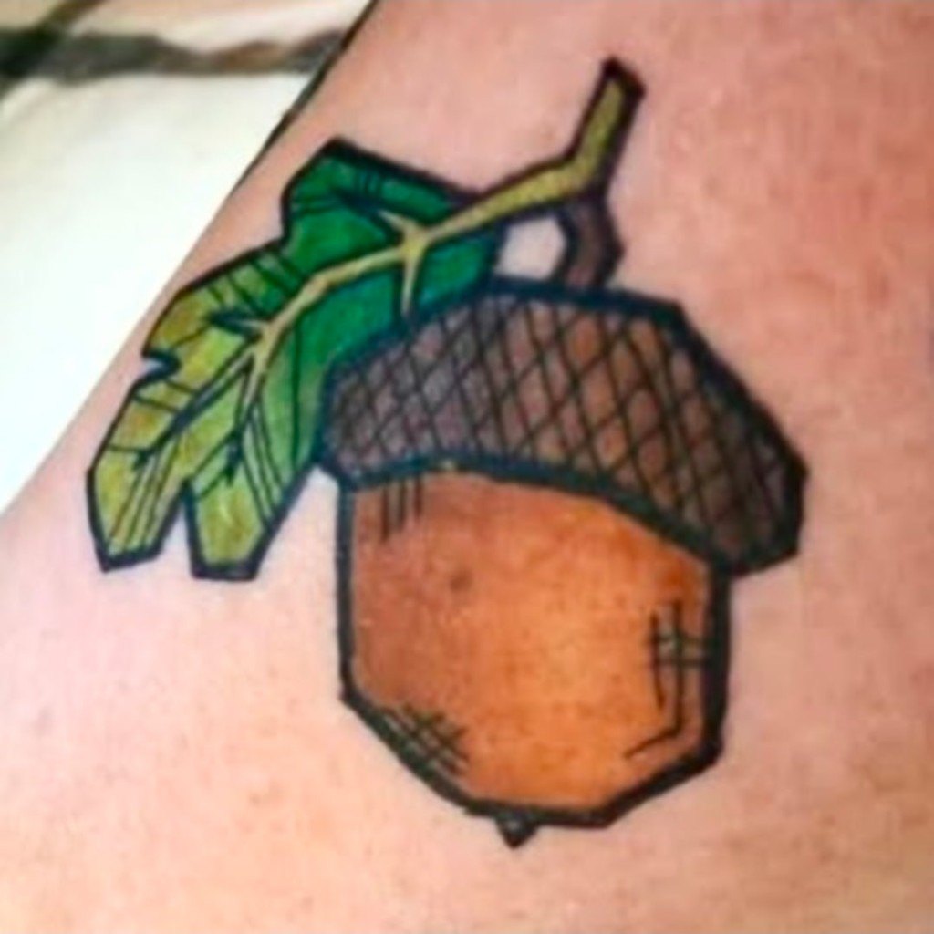 Illustrative acorn tattoo with a green oak leaf, bold outlines, and cross-hatch shading by Leni B. at Fargo Tattoo in Fargo, North Dakota
