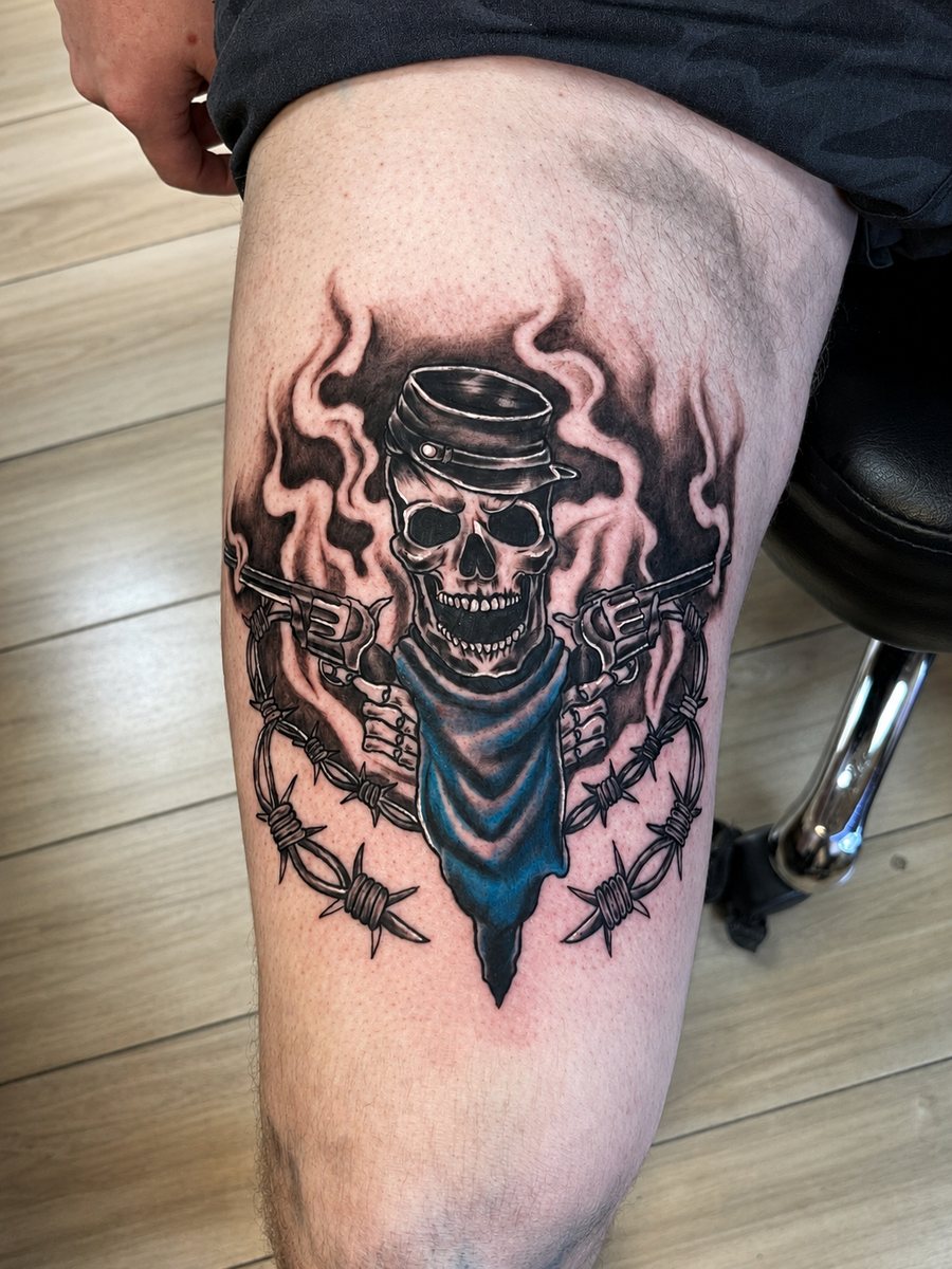 Skull in a cap holding revolvers, framed by barbed wire and blue fabric, tattooed by Shawn Lowe at Fargo Tattoo in Fargo, North Dakota